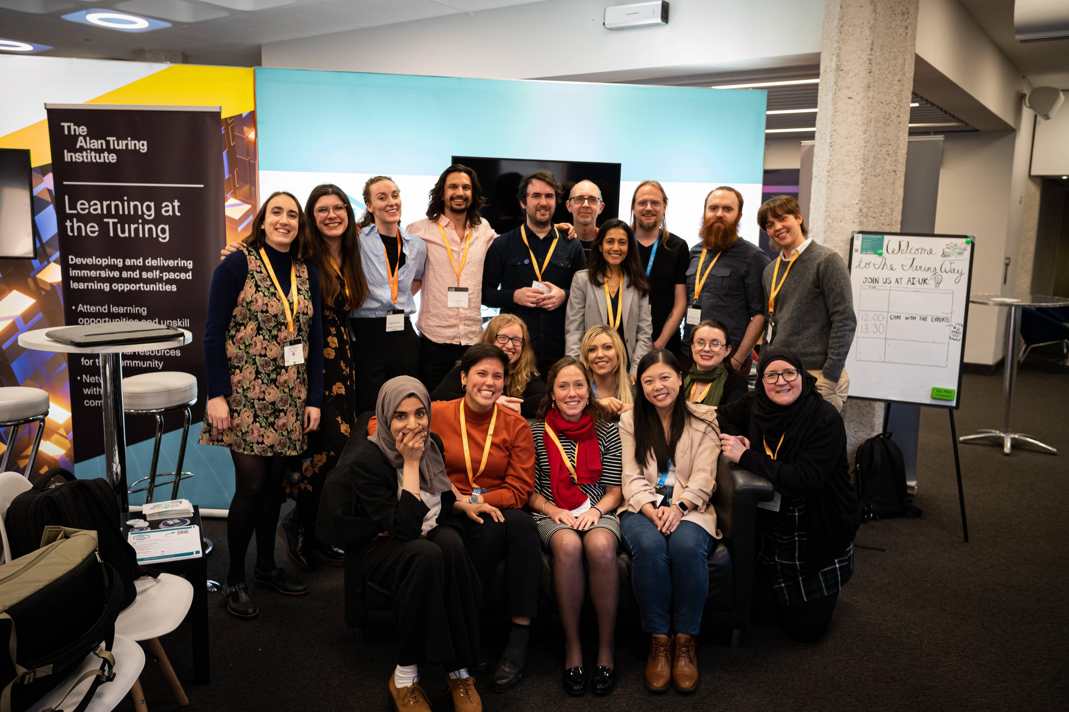 Alt: Photo of The Turing Way team members at AIUK in London on the 21 March. There are 18 people from the Alan Turing Institute in the picture, some sitting on a couch and some standing in front of The Turing Way stand