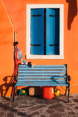 A window with blue wooden shutters on an orange wall. A blue bench below the window. Housekeeping objects laying around the bench.