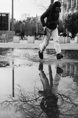 A monochrome photo. A woman runs through a puddle, with both feet above the water.