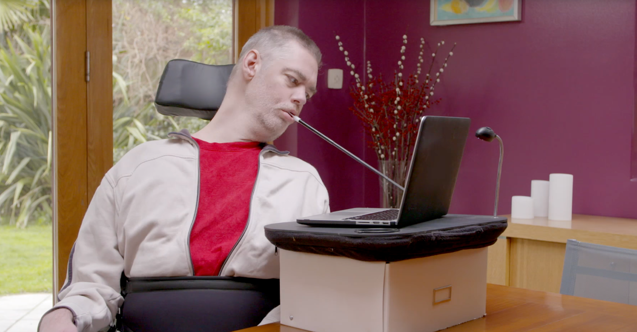 A man in a wheelchair is typing with a mouth stick (image source: W3C)