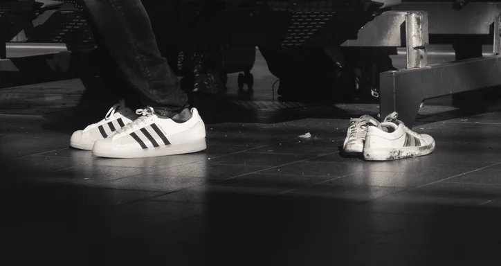 A black and white close-up photo of legs wearing brand-new sneakers. A similar pair of worn-out shoes lies nearby, leading to an interesting juxtaposition. The location is a train station.