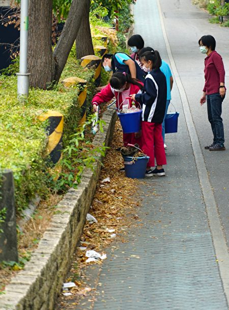  在河岸边道路上，带着夹子与水桶弯腰捡拾垃圾的学生与老师。