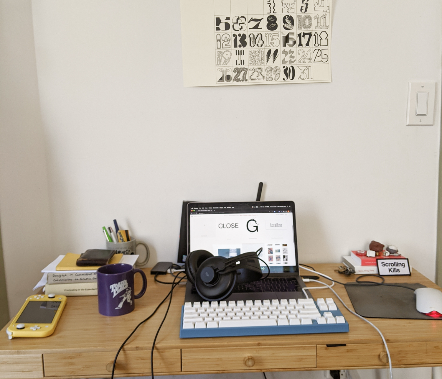 A desk workspace with a computer, keyboard, mouse and several books on it. A calendar hangs above.