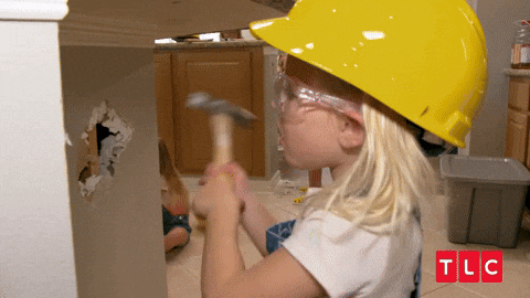 Image of a girl wearing a yellow construction hat and hitting a wall with a hammer