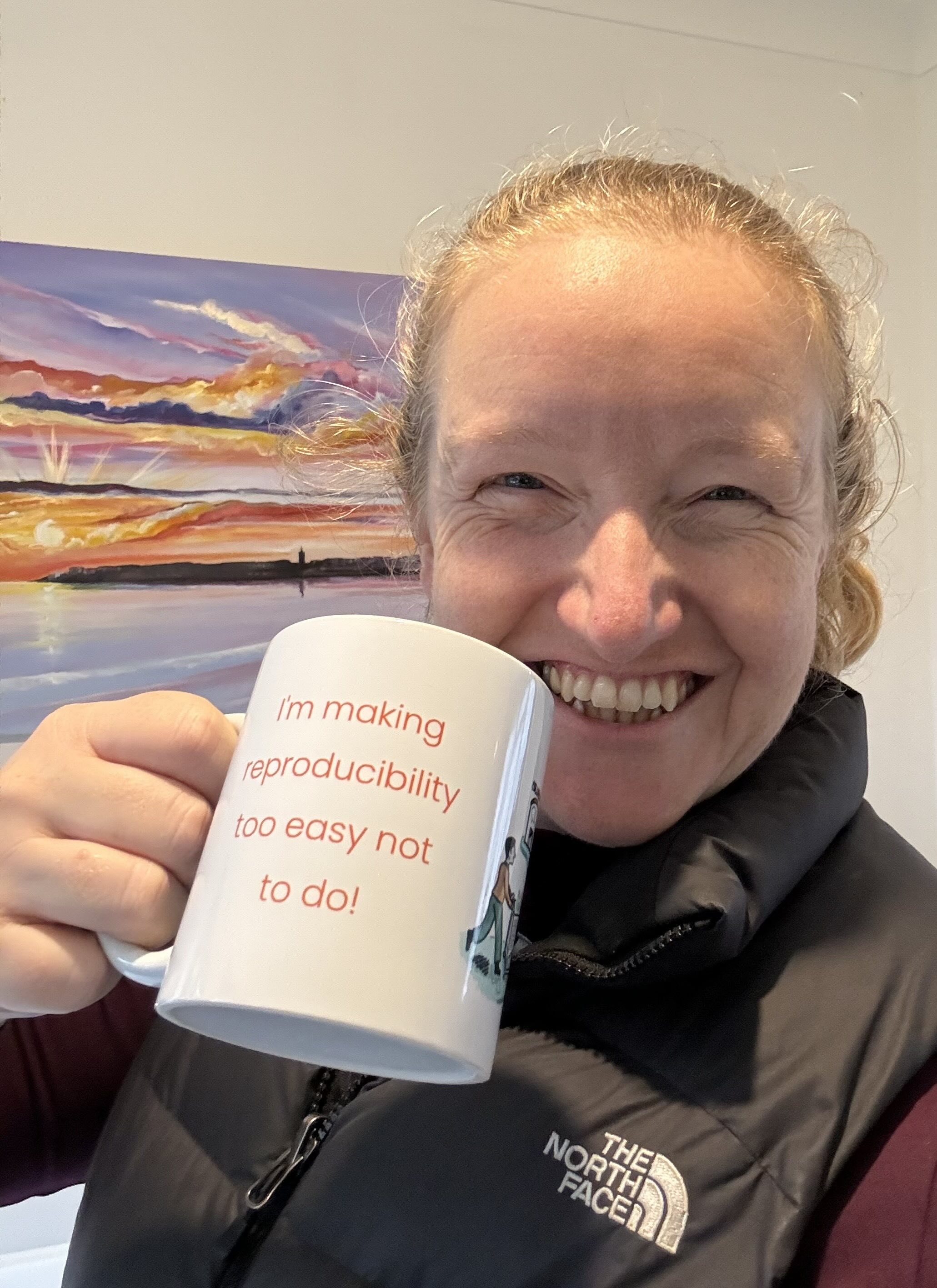 Alt: Kirstie, a blond white woman, taking a selfie holding the mug. The back - now showing to the camera - reads "I'm making reproducibility too easy not to do!". Behind her is a painting of a sunset.