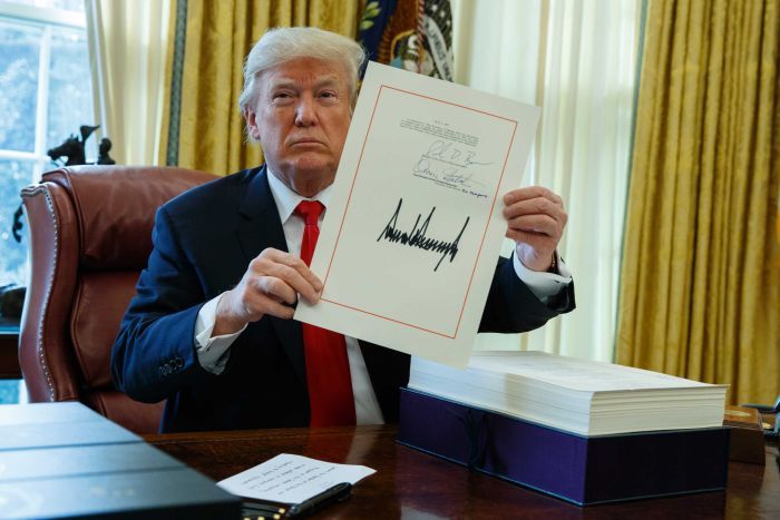 Donald Trump holds tax bill while sitting at his desk in the Oval Office with a serious look on his face