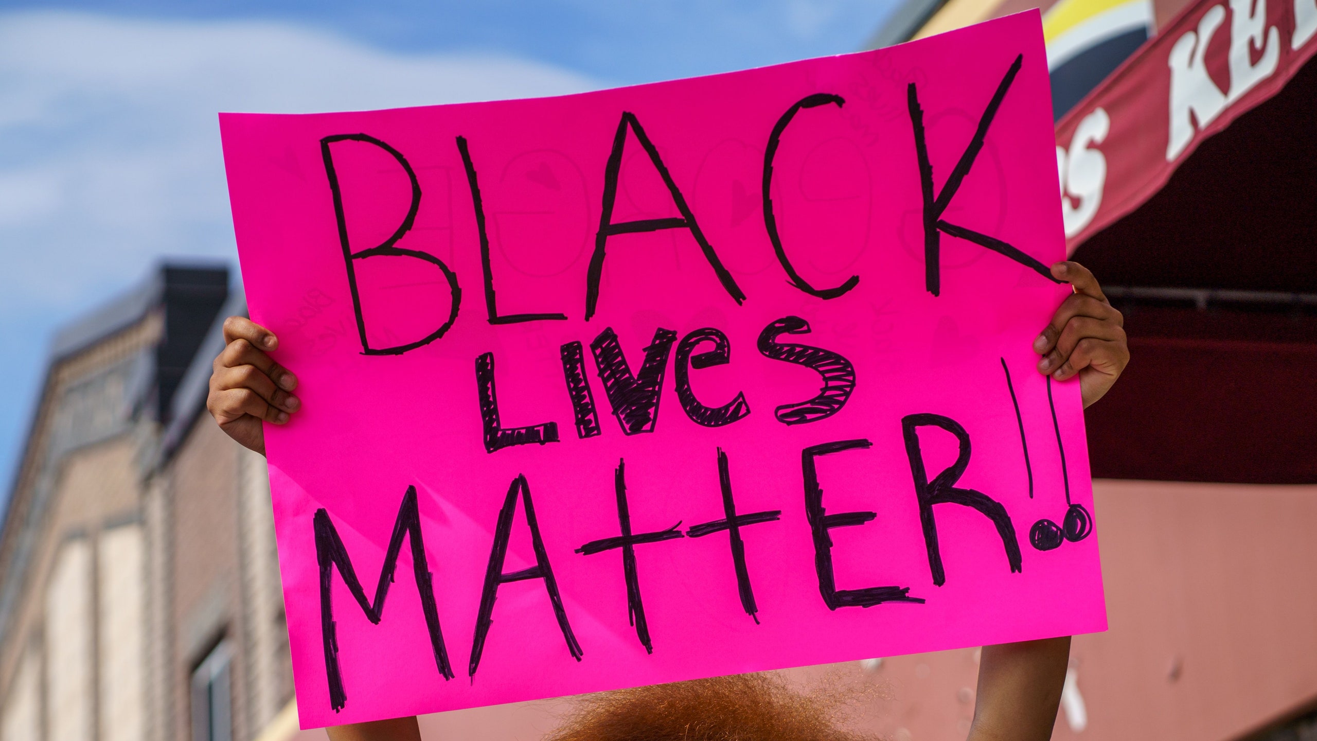 Image of a Black protester holding a pink sign reading in handwritten black lettering BLACK LIVES MATTER at a street protest