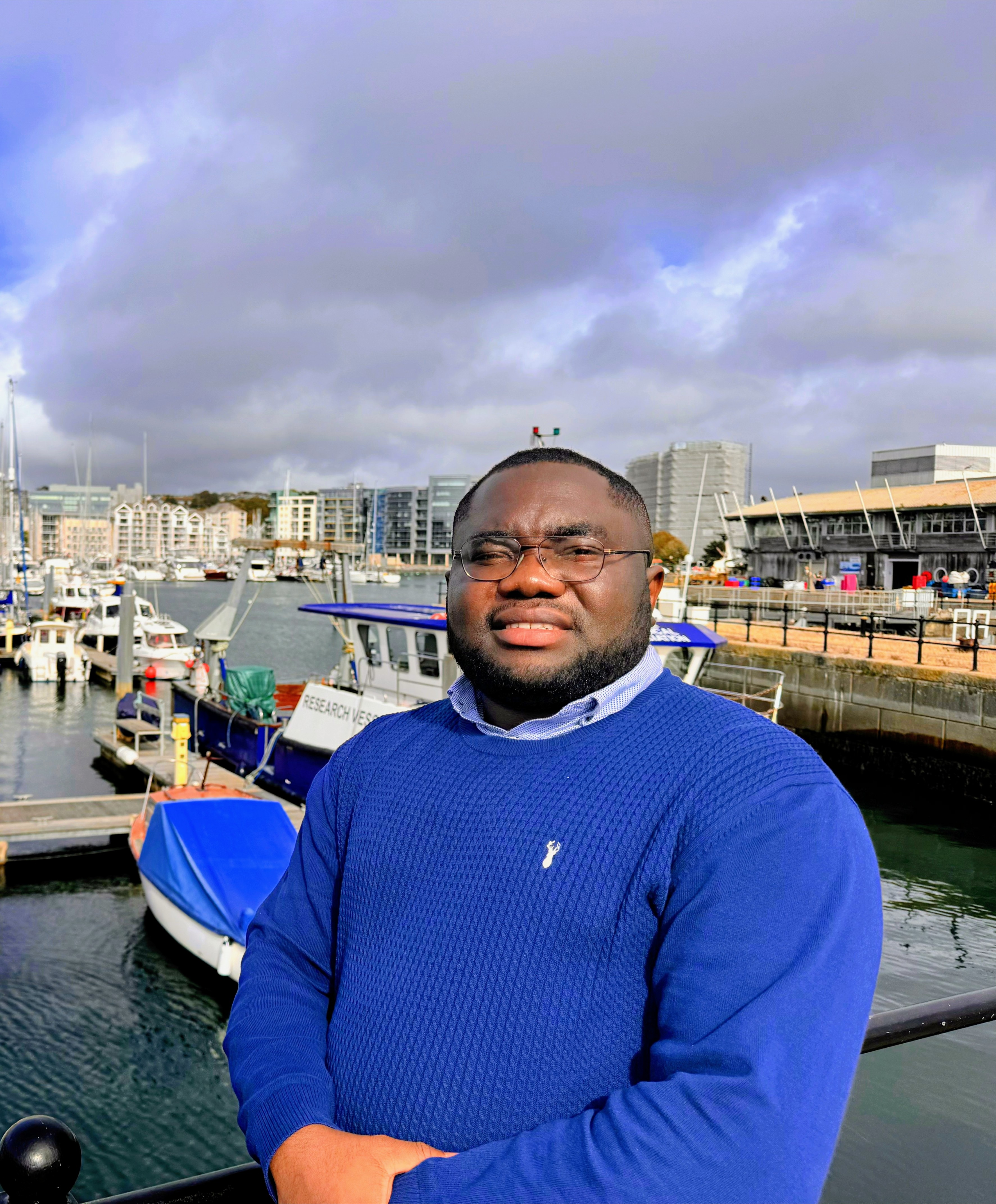 "Dr. Chidiebere Emmanuel Okechukwu, expert in echocardiography and public health, wearing a blue sweater and smiling at the Barbican waterfront in Plymouth, UK, with moored boats in the background"
