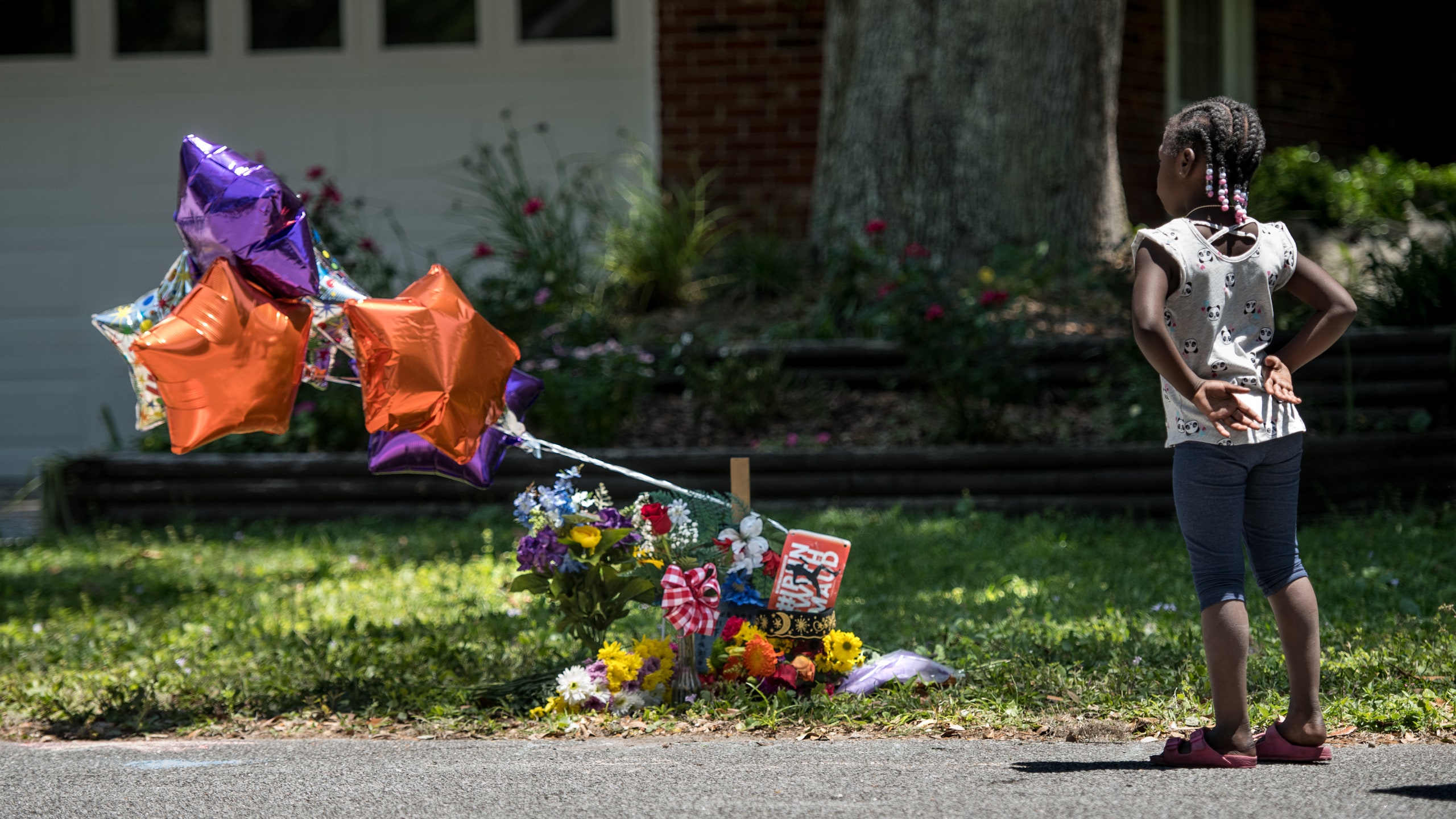 Image of a young black girl right of frame facing away from the camera as she considers a memorial of flowers and...