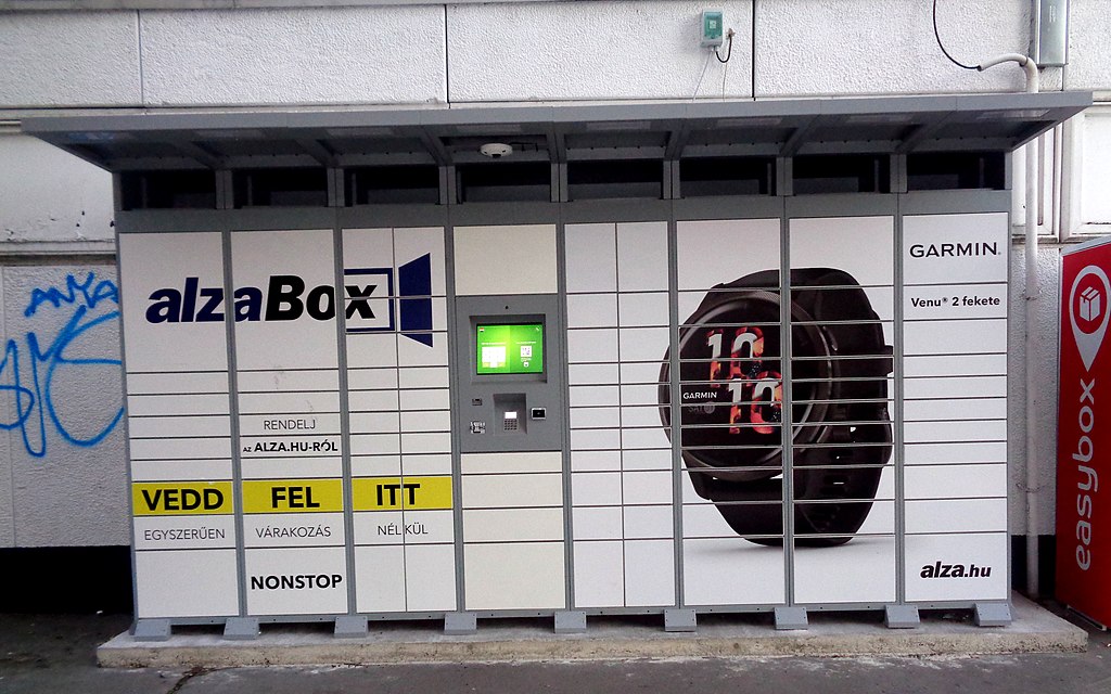 A large locker system labeled "alzaBox" with promotional graphics on its doors. It features a black Garmin Venu 2 watch advertisement on the front. The locker has a green interactive screen and keypad on one section. There is graffiti on the wall to the left and a sign labeled "easybox" on the right.