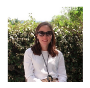 Alex has long brown hair and sunglasses. She is similing, and sitting with a long white sleeve shirt in front of a green bush