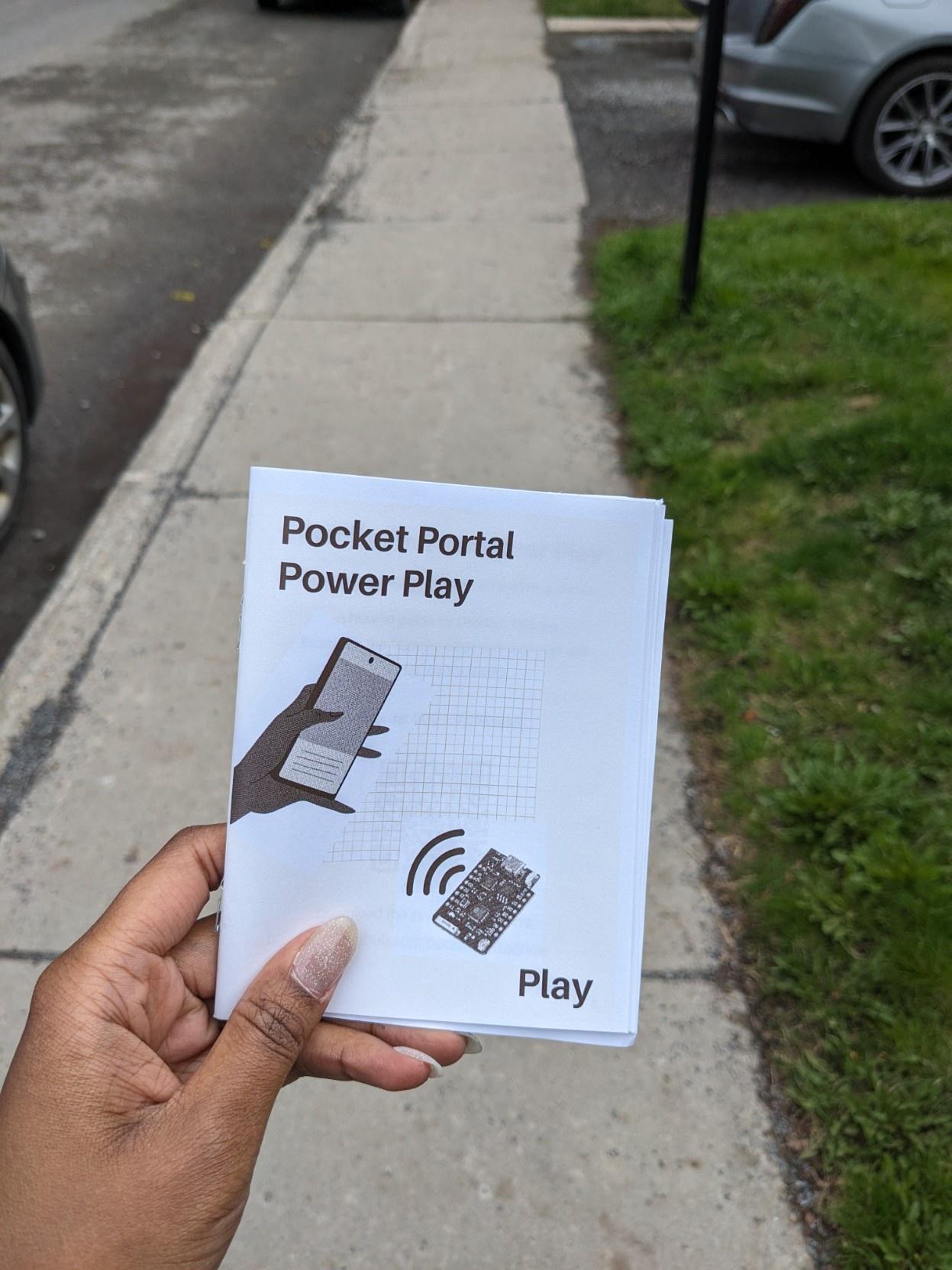 Picture of my hand holding the first print of the "Pocket Portal Power Play" zine. I'm standing outside, on a random sidewalk in Montréal, walking back from the print store where I feverishly sliced and stapled 20 zines for participants.