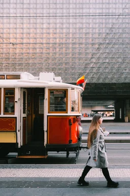 A woman walks on a street, next to a historic tram. A building with a glass square pattern in the background.