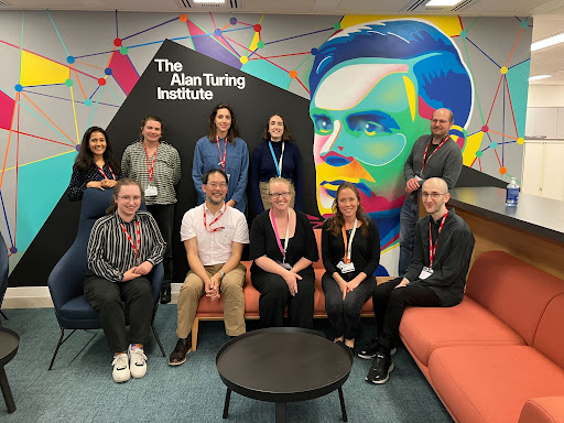 Alt: Picture of attendees of the Practitioners Hub workshop, taken at The Alan Turing office in London, on the back wall there is colourful painting of Alan Turing. Standing, from left to right: Malvika Sharan, Lucy Stephenson, Lauren Wool, Vicky Hellon and Brian Tarran. Sitting, from left to right: Rowan Hemsi, Neil Chue Hong, Kirstie Whitaker, Alexandra Araujo Alvarez and Raphael Sonabend