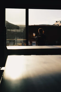 A waiter holding a tray with a glass enters a building. The photo is taken from the inside. The sun is shining outside. The waiter is mostly a silhouette, but the light highlights their red hair and the glass.