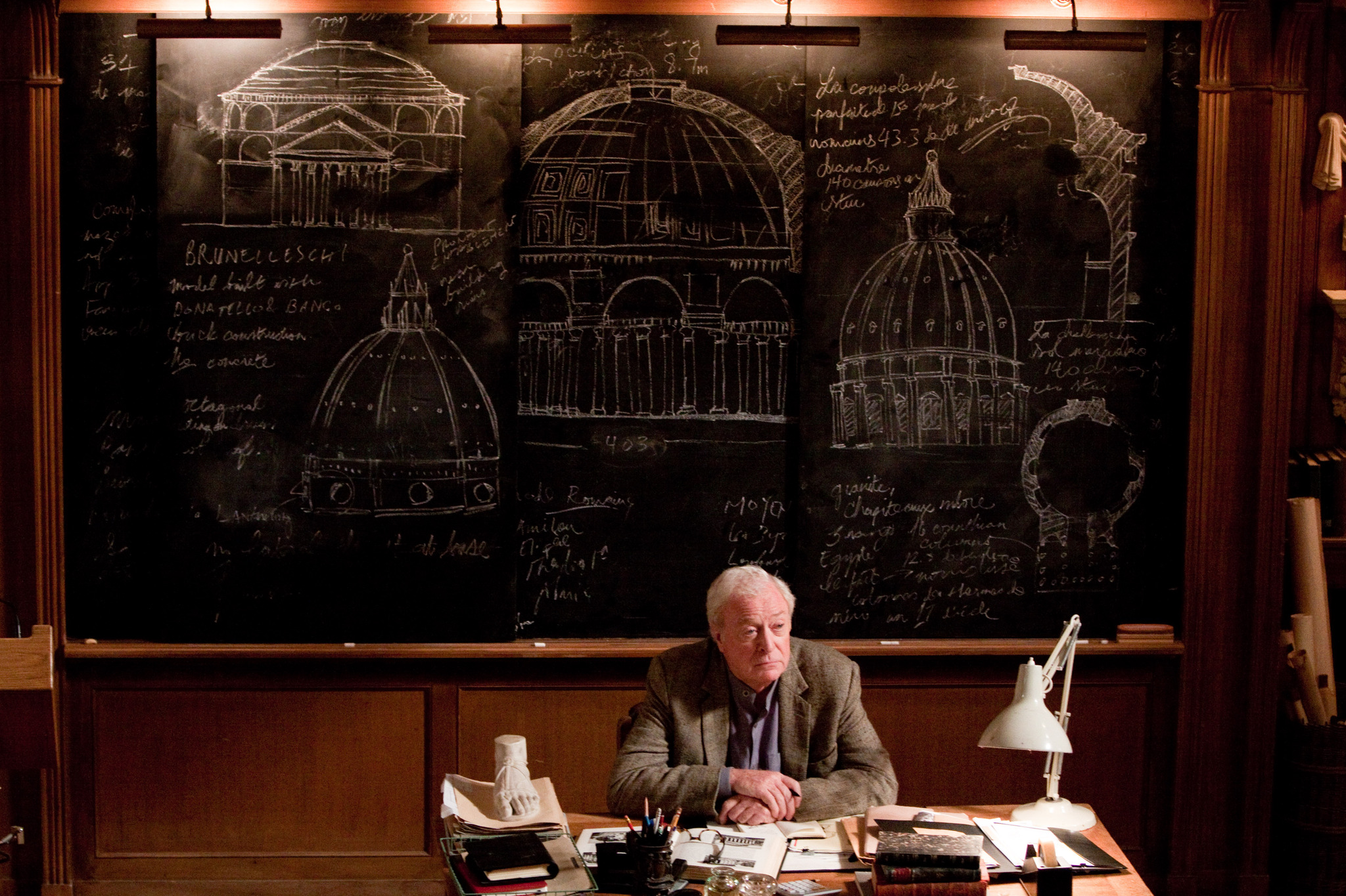 Michael Caine sitting at a desk in front of a chalkboard full of
        mathematical formulae and architectural drawings