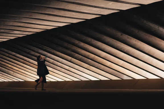 A person walking at night under a structure of lines, lights and shadows drinks from a bottle.
