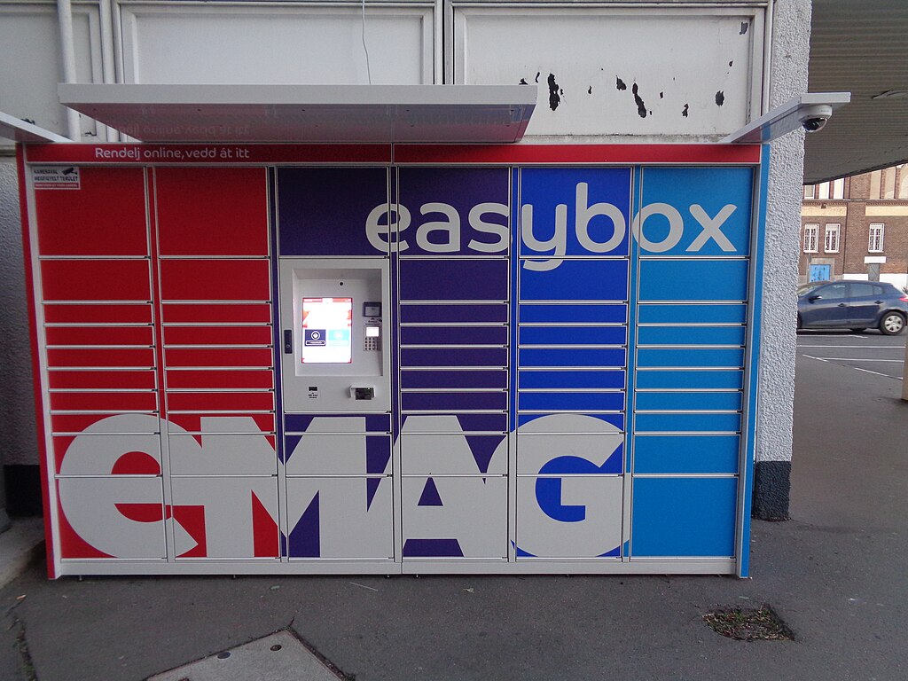 A red and blue eMAG easybox parcel locker is positioned on a sidewalk, featuring multiple compartments and a digital interface for package pickup. A car is visible in the background.