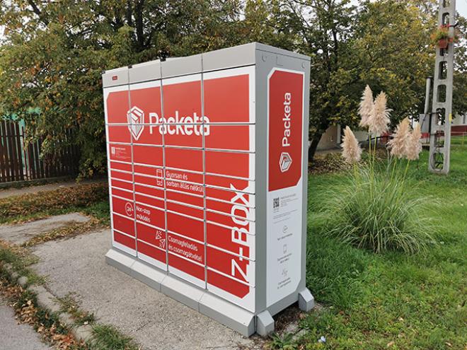 A red and white Packeta Z-BOX locker outdoors, surrounded by grass and trees. The locker is designed for parcel delivery and pickup.