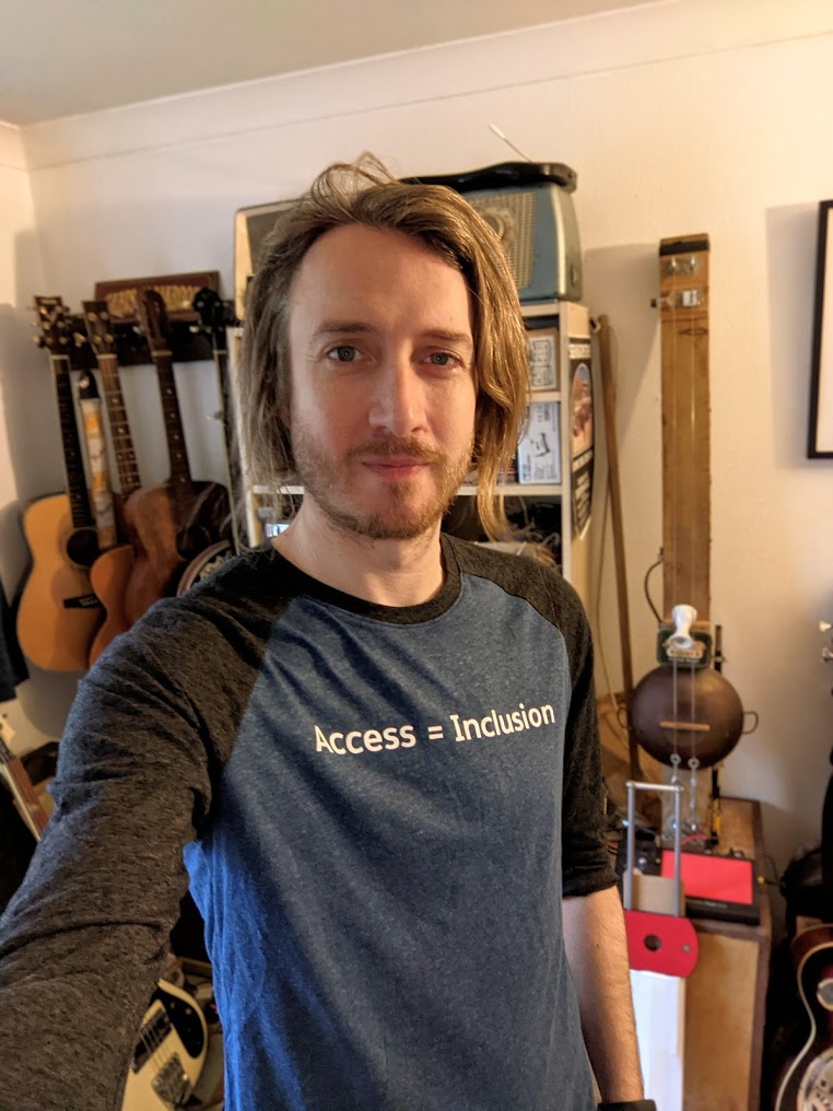 Mark in a blue t-shirt that read an access = inclustion.  in the backgroudn there are several guitars including a DIY 2 sting diddley bow.