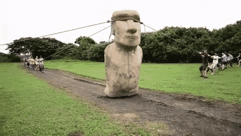 A moai statue being "walked" by several people holding a rope tied around its face at each side, rocking it from side to side to move it forwards.